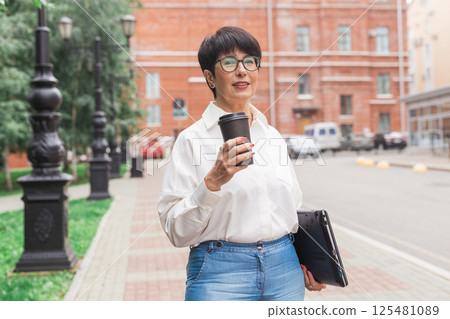Mature woman with coffee and laptop standing outdoors. Freelance work, career focus and independent lifestyle Mature woman with coffee and laptop standing outdoors. Freelance work, career focus and independent lifestyle 125481089