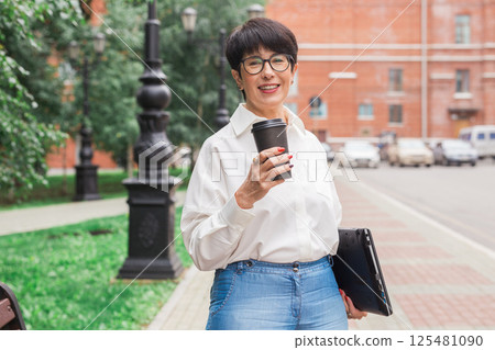 Senior woman enjoying coffee during walk with laptop. Urban lifestyle, freelance work and active aging 125481090