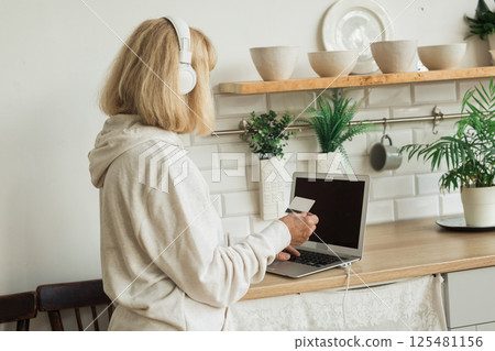 Senior woman making an online payment at home with laptop and headphones. Digital literacy and daily online activity in senior life 125481156