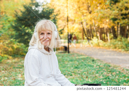 Senior woman walking with headphones in the park during fall, copy space. Active lifestyle, happiness, movement, freedom, and mindful aging in nature 125481244