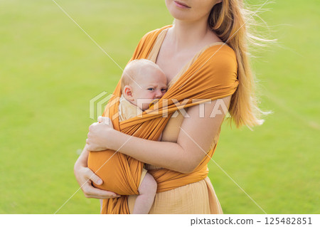 Mother holding her little baby in a yellow sling in the park. Warm and loving family moment. Babywearing, parenting, and mother-child bonding concept 125482851