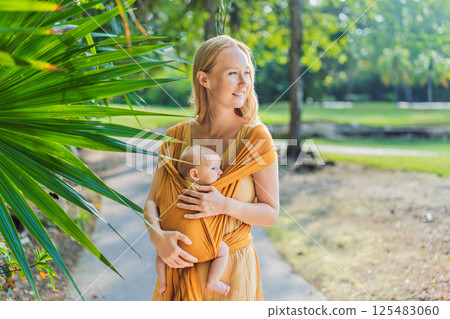 Mother holding her little baby in a yellow sling in the park. Warm and loving family moment. Babywearing, parenting, and mother-child bonding concept Mother holding her little baby in a yellow sling in the park. Warm and loving family moment. Babywearing, parenting, and mother-child bonding concept 125483060