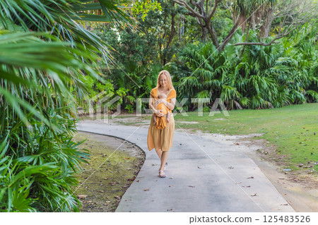 Mother holding her little baby in a yellow sling in the park. Warm and loving family moment. Babywearing, parenting, and mother-child bonding concept Mother holding her little baby in a yellow sling in the park. Warm and loving family moment. Babywearing, parenting, and mother-child bonding concept 125483526