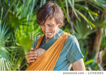 Father holding his little baby in a yellow sling in the park. Warm and loving family moment. Babywearing, fatherhood, and parent-child bonding concept 125483663