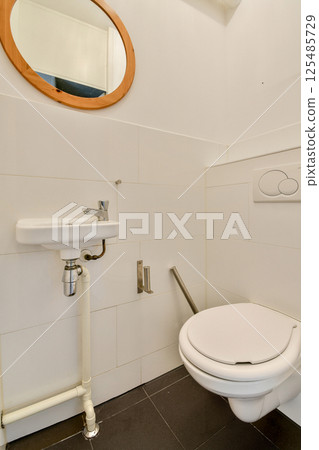 A sleek and minimalistic bathroom featuring a modern toilet, wall-mounted sink, and stylish circular mirror, emphasizing a contemporary aesthetic. A sleek and minimalistic bathroom featuring a modern toilet, wall-mounted sink, and stylish circular mirror, emphasizing a contemporary aesthetic. 125485729