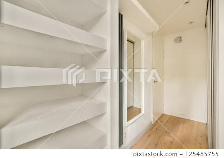 A clean and minimalist hallway featuring sleek white shelves and a mirror with natural wood flooring, creating an airy and spacious feel. 125485755