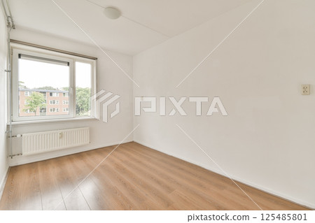A minimalistic empty room featuring a large window allowing natural light, with wooden flooring and white walls creating a spacious ambiance. A minimalistic empty room featuring a large window allowing natural light, with wooden flooring and white walls creating a spacious ambiance. 125485801