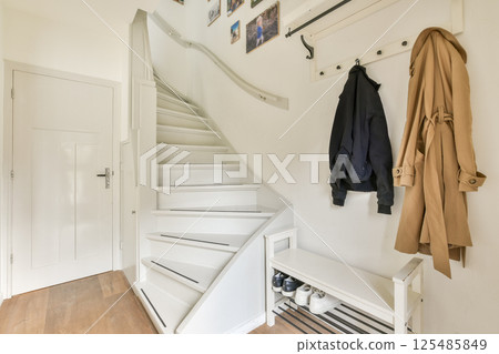 A clean and modern interior featuring a white staircase with natural light illuminating the space. Coats and shoes are neatly arranged, adding to the organized look. A clean and modern interior featuring a white staircase with natural light illuminating the space. Coats and shoes are neatly arranged, adding to the organized look. 125485849
