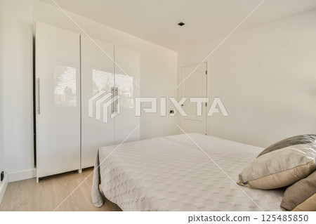 A bright and airy modern bedroom featuring a minimalist design with a large bed, sleek white wardrobes, and soft lighting elements for a tranquil ambiance. 125485850