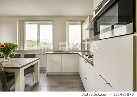 A stylish modern kitchen featuring sleek cabinetry, a wooden dining table, and bright windows that enhance the space with natural light. 125485876