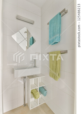 A sleek modern bathroom featuring a minimalist design with a stylish sink, mirrored wall, and vibrant towels hanging on racks. A small shelf holds additional towels. 125486213