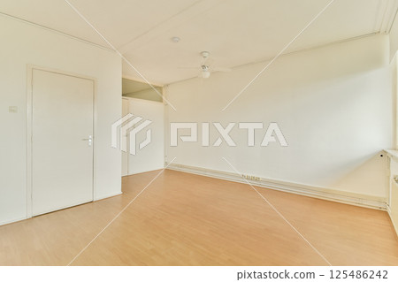 A simple and clean interior room showcasing light-colored walls and wooden flooring, providing a spacious and airy atmosphere. 125486242