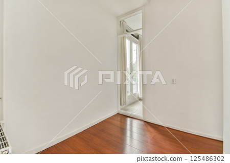 A bright and airy interior hallway with sleek wooden flooring and clean, white walls leading to a large window. The space reflects modern minimalist design principles. 125486302