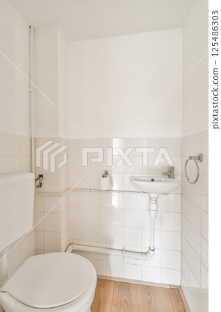 A modern bathroom featuring a minimalistic design with white tiled walls, a toilet, and a sink. The wooden floor adds warmth to the overall look. 125486303