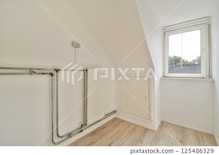 An interior view showcasing minimalistic design with white walls, wooden flooring, and a window providing natural light. Industrial piping adds a unique touch to the space. 125486329