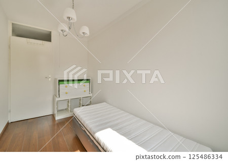 A bright and minimalist bedroom featuring a single mattress on a wooden floor, complemented by a modern chandelier and a compact storage unit. 125486334