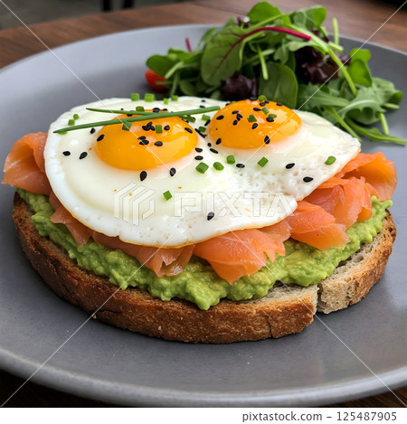 A slice of avocado toast is topped with smoked salmon and two fried eggs, served with a small salad A slice of avocado toast is topped with smoked salmon and two fried eggs, served with a small salad 125487905