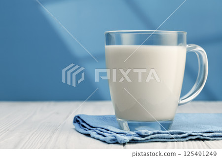Glass mug brimming with fresh milk, resting on blue napkin atop white wooden table Glass mug brimming with fresh milk, resting on blue napkin atop white wooden table 125491249