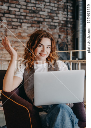 A young woman with curly red hair sits in a cafe with her laptop, looking confidently at the camera. Confident woman, modern entrepreneur, startup mindset, driven professional... 125495839