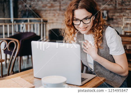 A young woman with curly red hair smiles while engaging in a video call from a cafe. Remote work and digital collaboration, hybrid workspace, online meeting, flexible work... A young woman with curly red hair smiles while engaging in a video call from a cafe. Remote work and digital collaboration, hybrid workspace, online meeting, flexible work... 125495847