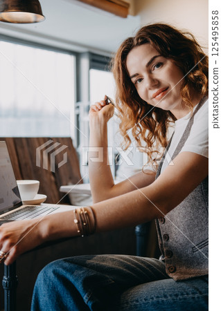 A young woman with curly red hair pauses work to smile at the camera in a cozy cafe setting. Women in digital professions, female freelancer, tech industry woman, creative remote worker... 125495858