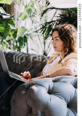 Young woman with auburn curly hair working on a laptop in a stylish indoor setting with plants. Remote work, digital nomad lifestyle, flexible work, location independence... 125495859