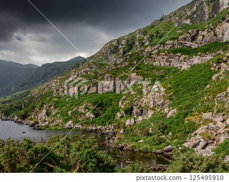 Overlooking a tranquil river, the image captures steep rocky hills blanketed in greenery. Dark clouds loom overhead, adding drama to the peaceful setting. 125495910