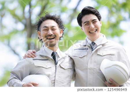 Image of a worker in construction management, etc., wearing work clothes and looking up in front of a building with fresh greenery in the background, looking at the camera with his shoulders crossed 125496949