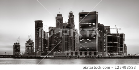 Monochrome city skyline with tall skyscrapers reflecting on calm water under a cloudy sky, Dubai Marina bay UAE 125498558