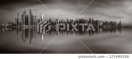 Monochrome city skyline with tall skyscrapers reflecting on calm water under a cloudy sky, Dubai Marina bay UAE 125498598