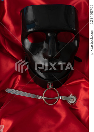 Adult sex games. Kinky lifestyle. Wartenberg Wheel and black mask on the red linen. Bdsm outfit Adult sex games. Kinky lifestyle. Wartenberg Wheel and black mask on the red linen. Bdsm outfit 125498792