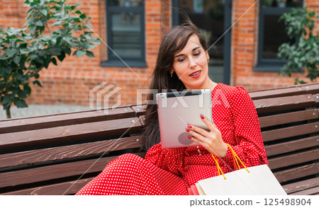 Woman sitting on bench with shopping bags and tablet. Digital lifestyle and modern shopping experience. 125498904