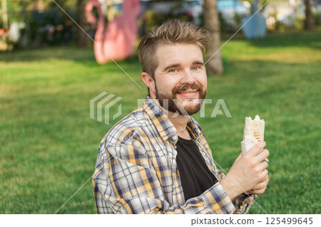 Smiling man holding a sandwich wrap while enjoying outdoor lunch. Concept of happiness, casual street food culture, and joyful moments during a break. 125499645