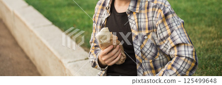 Man holding a street food wrap in craft paper outdoors. Concept of fast food, casual lifestyle, and outdoor snacking in urban park settings. Banner with copy space 125499648