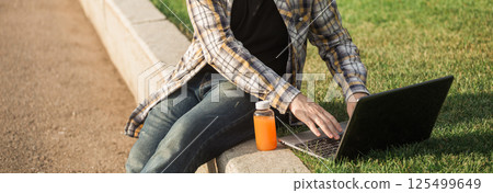 Man working outdoors on a laptop with juice beside him. Concept of remote work, productivity, and casual freelance lifestyle in urban parks. Banner with copy space 125499649