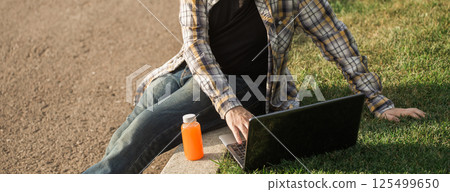 Man working outdoors on a laptop with juice beside him. Concept of remote work, productivity, and casual freelance lifestyle in urban parks. Banner with copy space 125499650