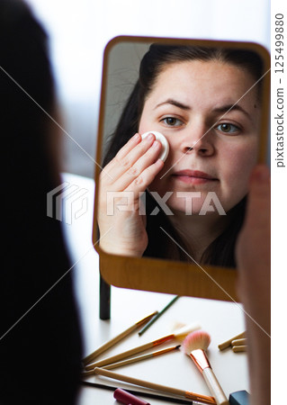 Vertical image of Millennial Caucasian woman removing make up from face while looking in mirror. Reflection in mirror. Getting ready for bed. Self care Beauty concept. Vertical image of Millennial Caucasian woman removing make up from face while looking in mirror. Reflection in mirror. Getting ready for bed. Self care Beauty concept. 125499880