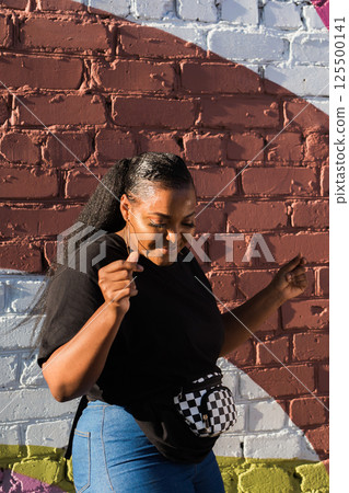 African american woman dancing near colorful brick wall during sunny day. Concept of joy, freedom, youth culture and positive urban lifestyle. African american woman dancing near colorful brick wall during sunny day. Concept of joy, freedom, youth culture and positive urban lifestyle. 125500141