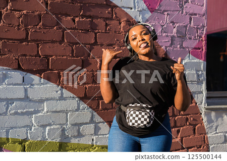 African american woman dancing playfully with headphones against painted wall. Concept of joy, energy, fun and youth lifestyle. African american woman dancing playfully with headphones against painted wall. Concept of joy, energy, fun and youth lifestyle. 125500144