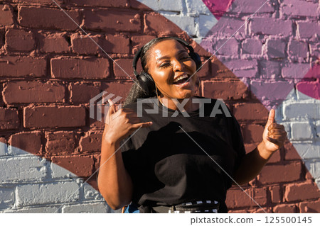African american woman dancing playfully with headphones against painted wall. Concept of joy, energy, fun and youth lifestyle. 125500145