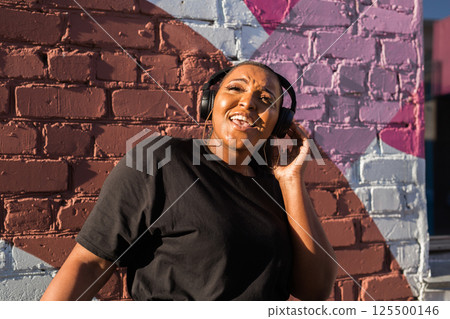 African american woman dancing playfully with headphones against painted wall. Concept of joy, energy, fun and youth lifestyle. 125500146