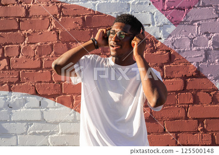 Young african american man dancing with headphones on colorful wall background. Concept of music, joy, freedom and urban lifestyle. Copy space Young african american man dancing with headphones on colorful wall background. Concept of music, joy, freedom and urban lifestyle. Copy space 125500148
