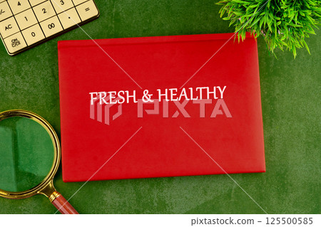 A vibrant red folder titled fresh and healthy is placed on a textured green surface. A magnifying glass and a small plant add to the composition, inviting thoughts of wellness and nutrition 125500585
