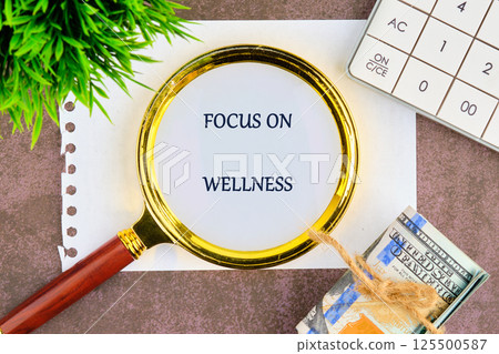 A magnifying glass highlights the phrase focus on wellness placed on a surface with a small plant and nearby items including notes and currency that symbolize self-care and financial wellness 125500587
