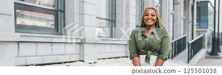 Confident african american woman standing against city architecture smiling. Concept of positivity, urban lifestyle and self-confidence. Banner with copy space 125501038