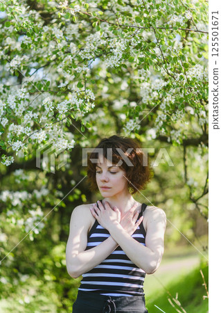 Woman standing under blooming tree, touching chest with hands. Woman standing under blooming tree, touching chest with hands. 125501671