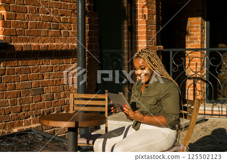 African american woman smiling and holding a tablet while sitting on outdoor cafe. Communication, happiness, and digital connection with loved ones. Banner with copy space 125502123