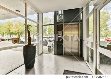 A contemporary building entrance showcasing an elevator and large windows, surrounded by greenery and parked cars. It illustrates a blend of architecture and nature. 125502358