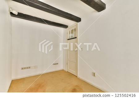 A minimalistic interior room featuring white walls, wooden beams, and a simple door, offering a clean and spacious atmosphere. 125502416