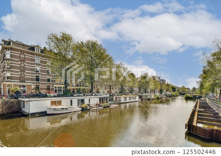 A picturesque view of a serene canal lined with lush trees and charming boats, reflecting the beauty of nature and urban life in harmony. A picturesque view of a serene canal lined with lush trees and charming boats, reflecting the beauty of nature and urban life in harmony. 125502446